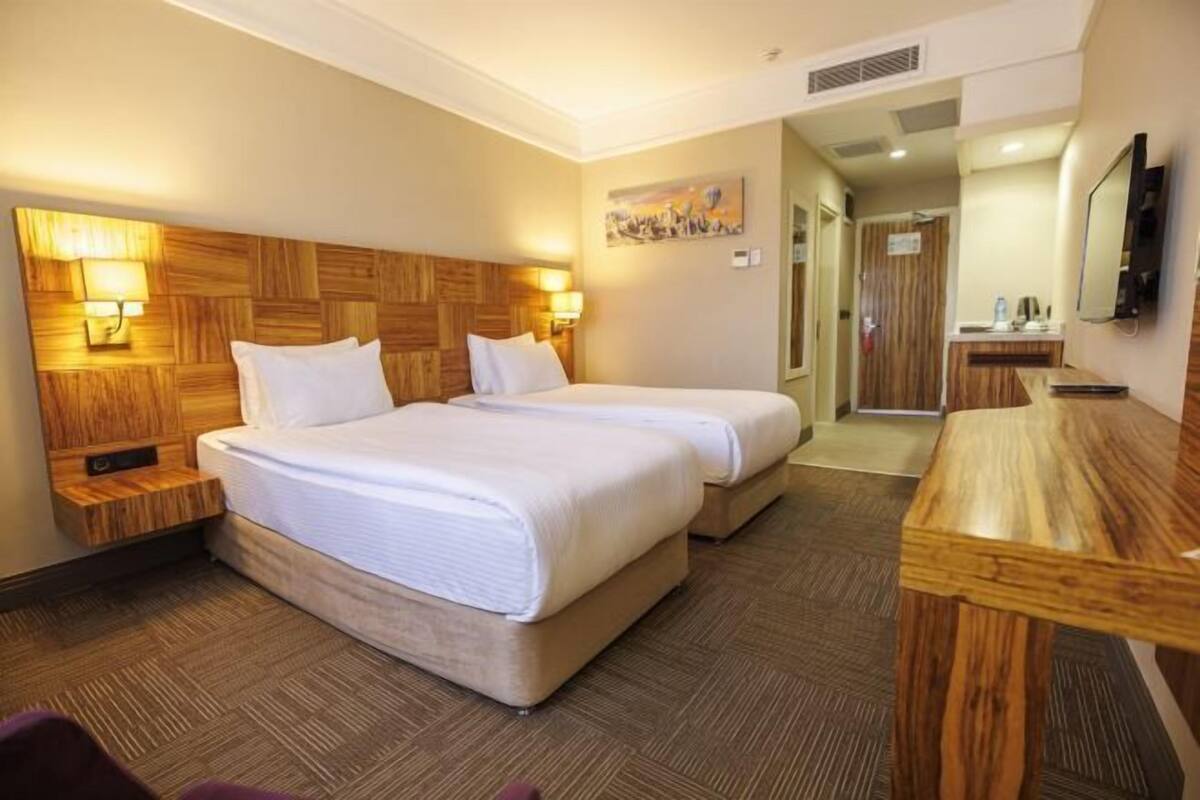 Ein Hotelzimmer mit zwei Betten, einem Holz-Kopfteil, Nachttischlampen, einem Holztisch und einem Gemälde an der Wand.
