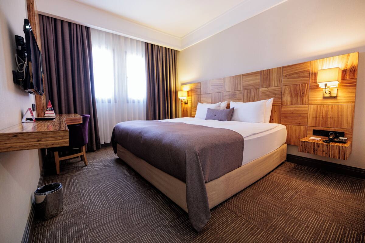Ein Hotelzimmer mit einem großen Bett, einem Schreibtisch mit Stuhl, einem Fernseher und einem hölzernen Bettkopfschild mit Wandleuchten.