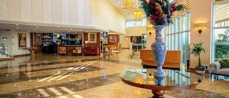 A spacious lobby with a polished marble floor, a reception desk, a coffee bar, and a decorative vase with dried flowers.