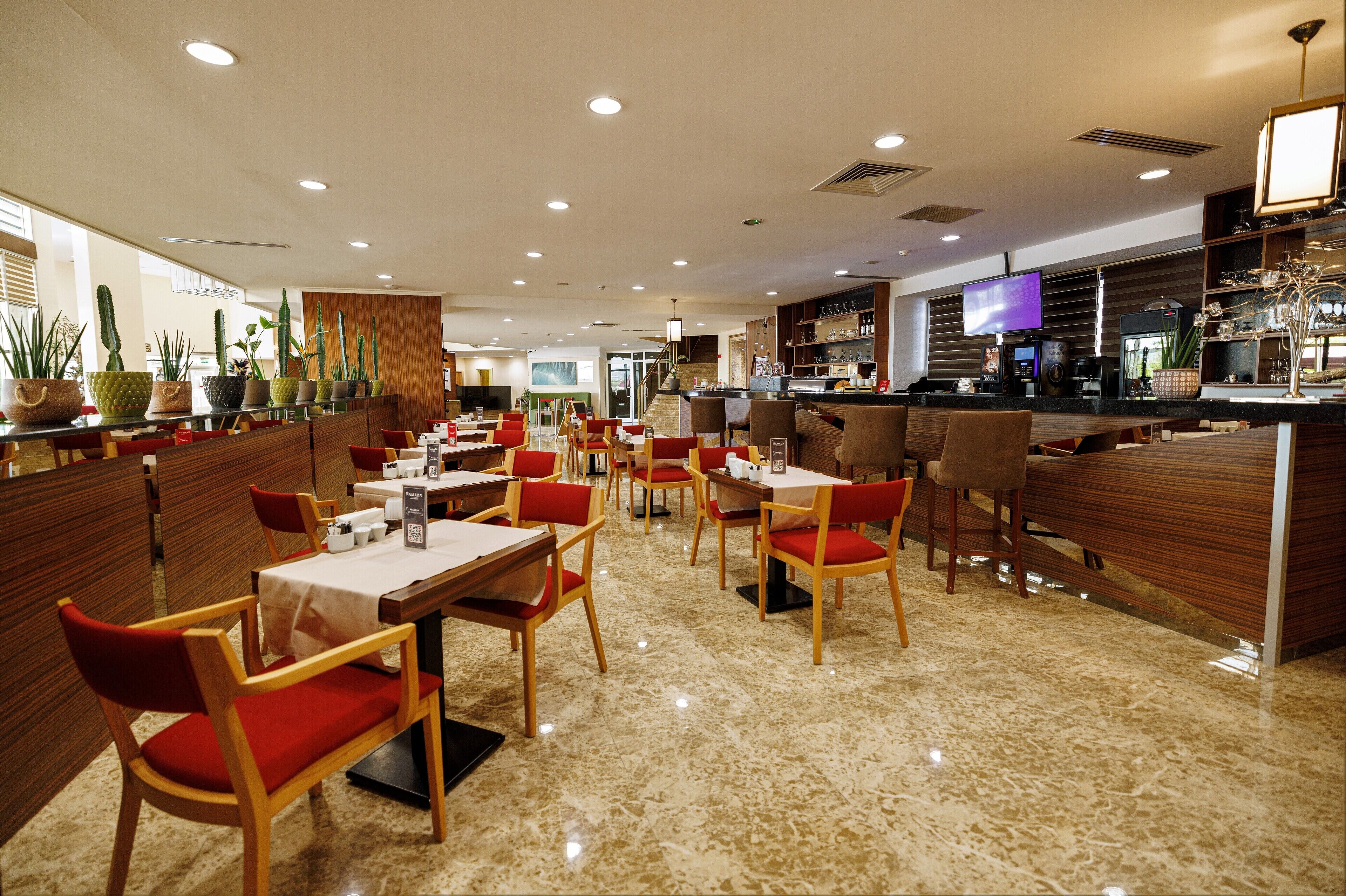 Un restaurante moderno con paneles de madera, sillas rojas, suelo de mármol y una zona de bar con televisor.