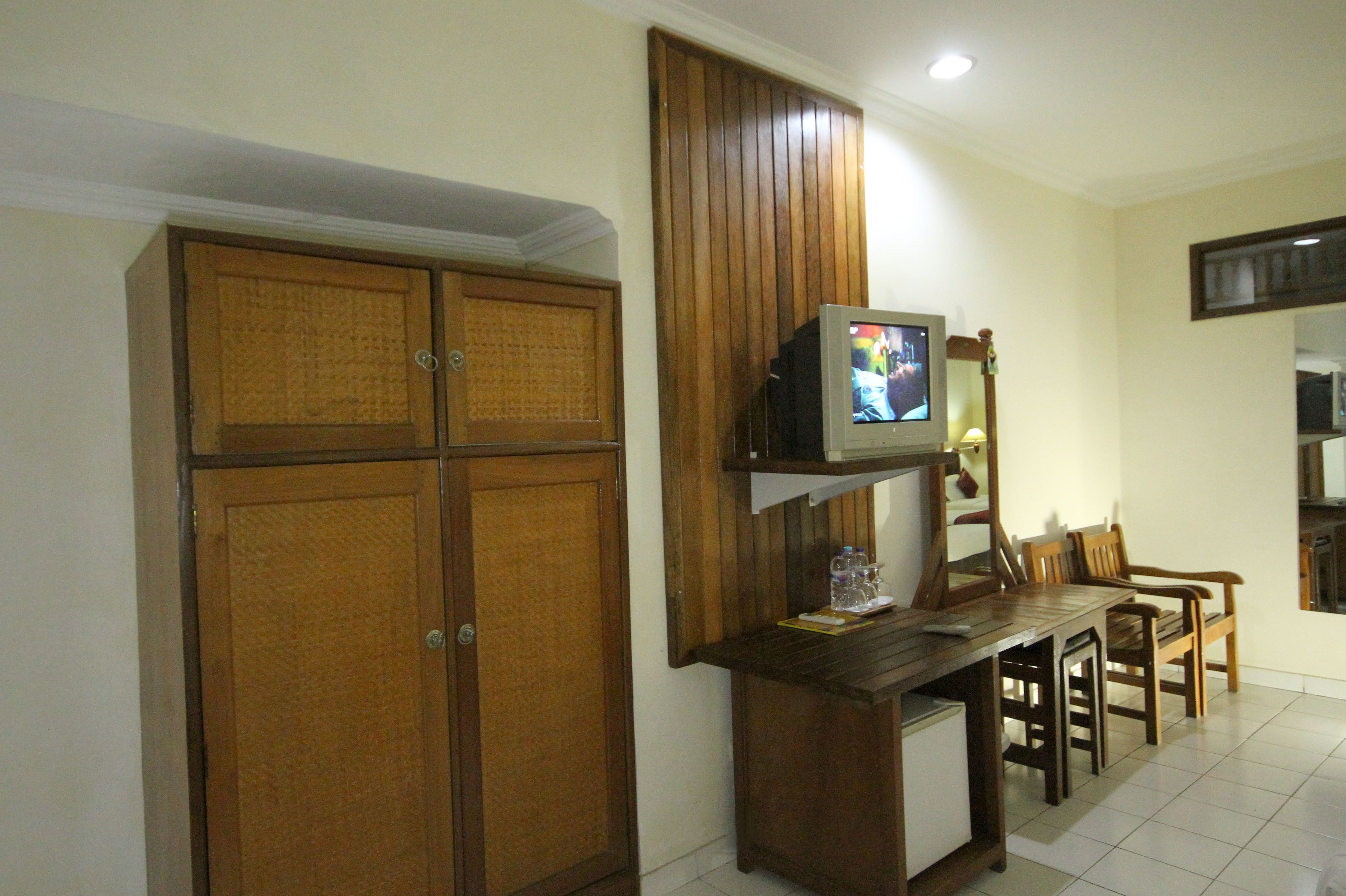 A room with a wooden wardrobe, a small TV on a shelf, and a dining table with chairs.