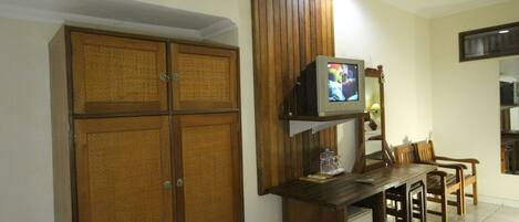 A room with a wooden wardrobe, a small TV on a shelf, and a dining table with chairs.