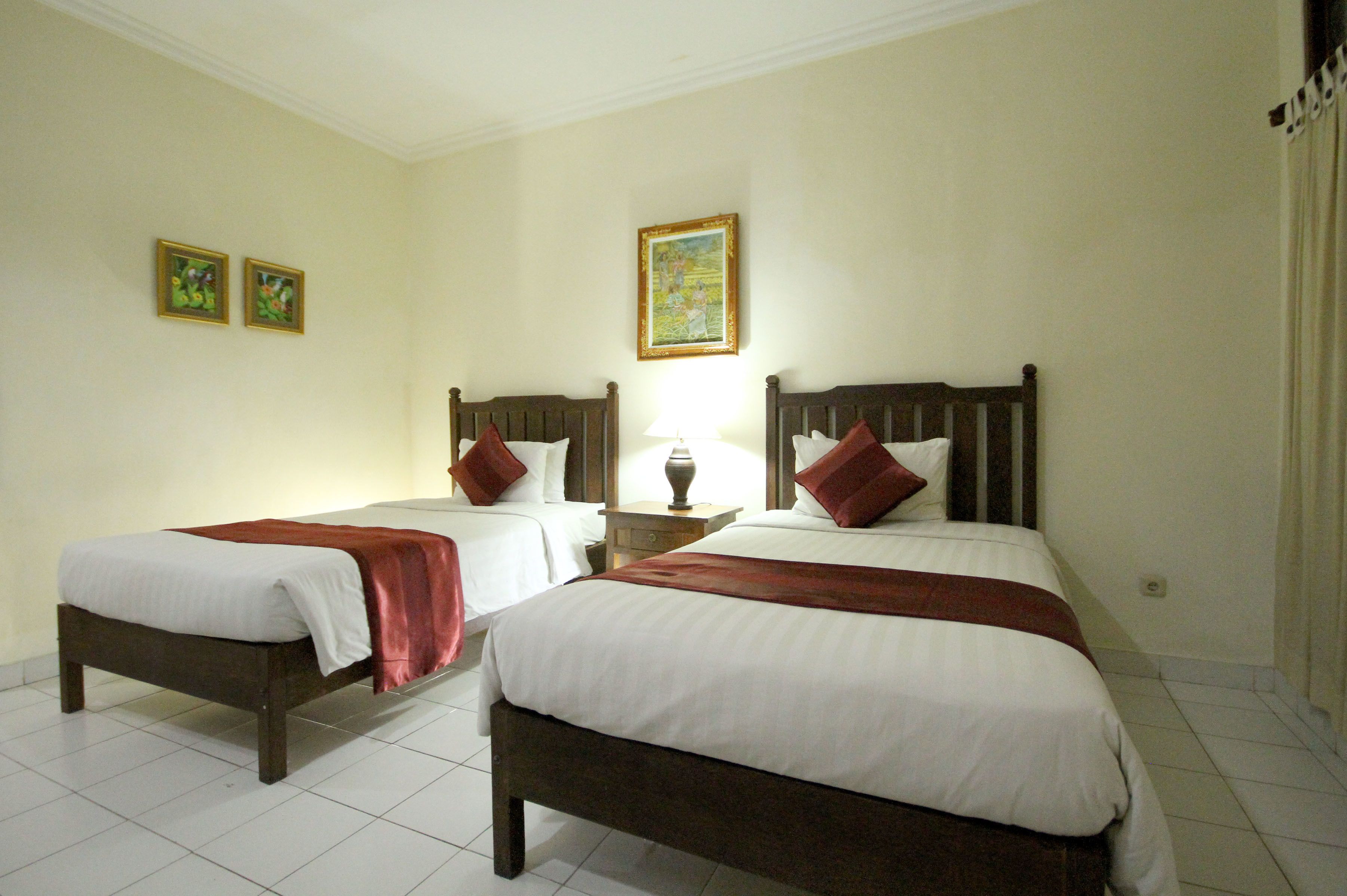 A hotel room with two single beds, a nightstand with a lamp, and two framed pictures on the wall.