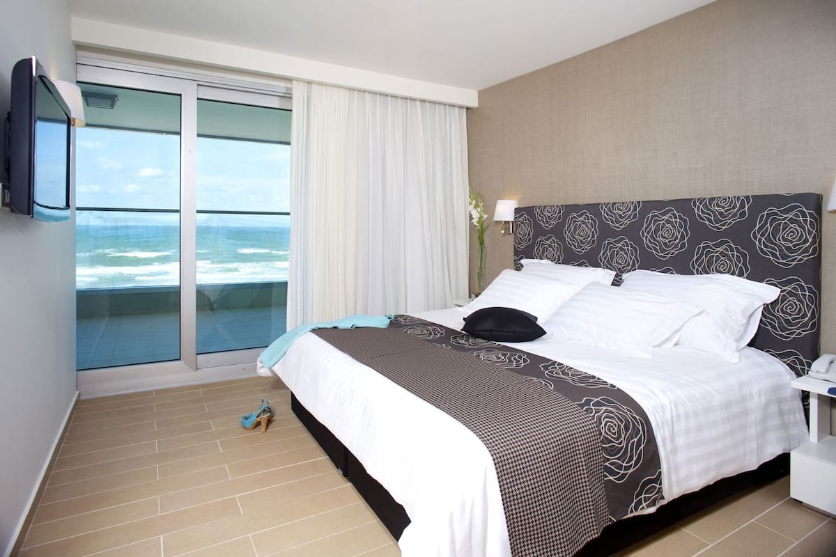 A hotel room with a bed, a TV, a nightstand, a lamp, and a view of the ocean.