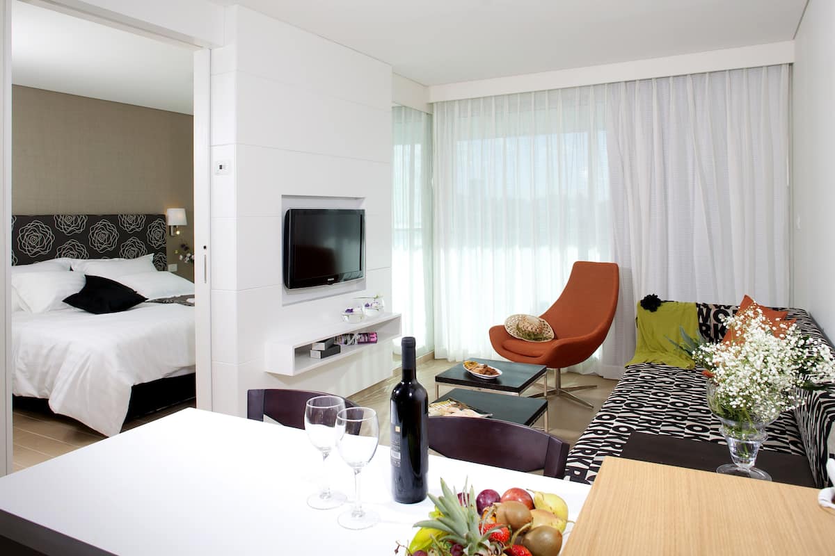 A hotel room with a bed, a TV, a dining table with wine glasses, a fruit bowl, and a sofa with a vase of flowers.