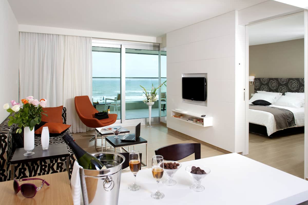 A modern hotel room with a balcony, a dining area with a table set for two, and a bedroom with a large bed and a TV mounted on the wall.