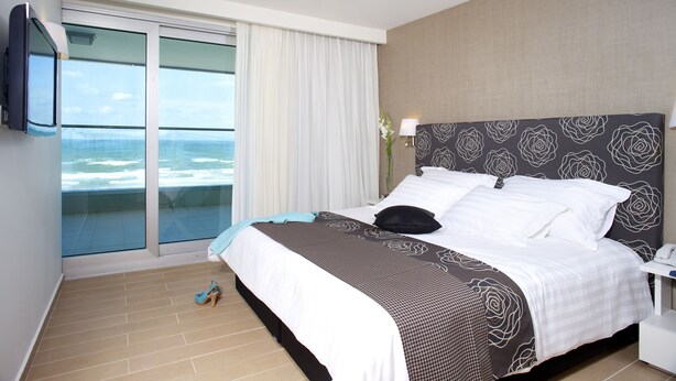 A hotel room with a bed, a TV, a nightstand, a lamp, and a view of the ocean.