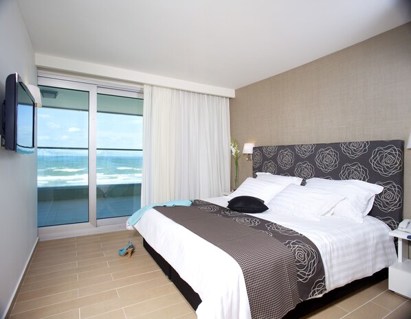 A hotel room with a bed, a TV, a nightstand, a lamp, and a view of the ocean. - Brown Seaside Ashdod (Ashdod)