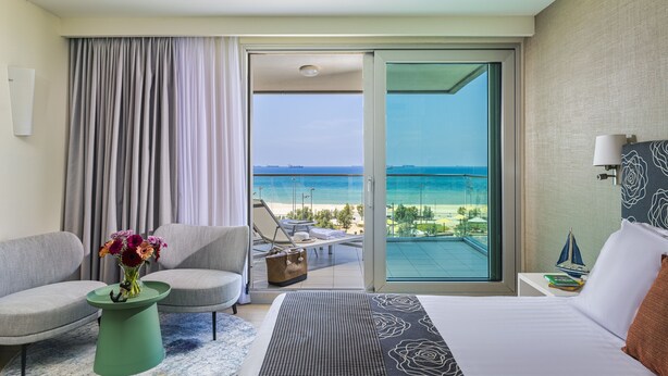 A hotel room with a bed, a chair, a small table with a vase of flowers, a view of the beach and ocean through a sliding glass door. - Brown Seaside Ashdod (Ashdod)