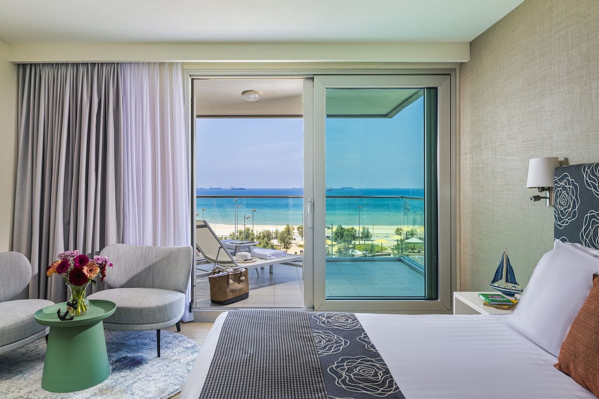A hotel room with a bed, a chair, a small table with a vase of flowers, a view of the beach and ocean through a sliding glass door.