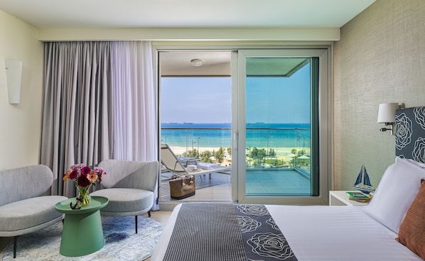 A hotel room with a bed, a chair, a small table with a vase of flowers, a view of the beach and ocean through a sliding glass door. - Brown Seaside Ashdod (Ashdod)