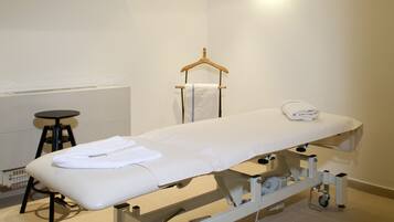 A clinical examination table with a white mattress, a stool, a white mat, and a wall-mounted shelf with two white lanterns.