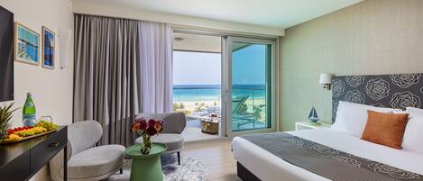 Habitación de hotel con cama, escritorio, silla, mesita con un jarrón de flores y vista a la playa a través de una puerta corrediza de cristal.