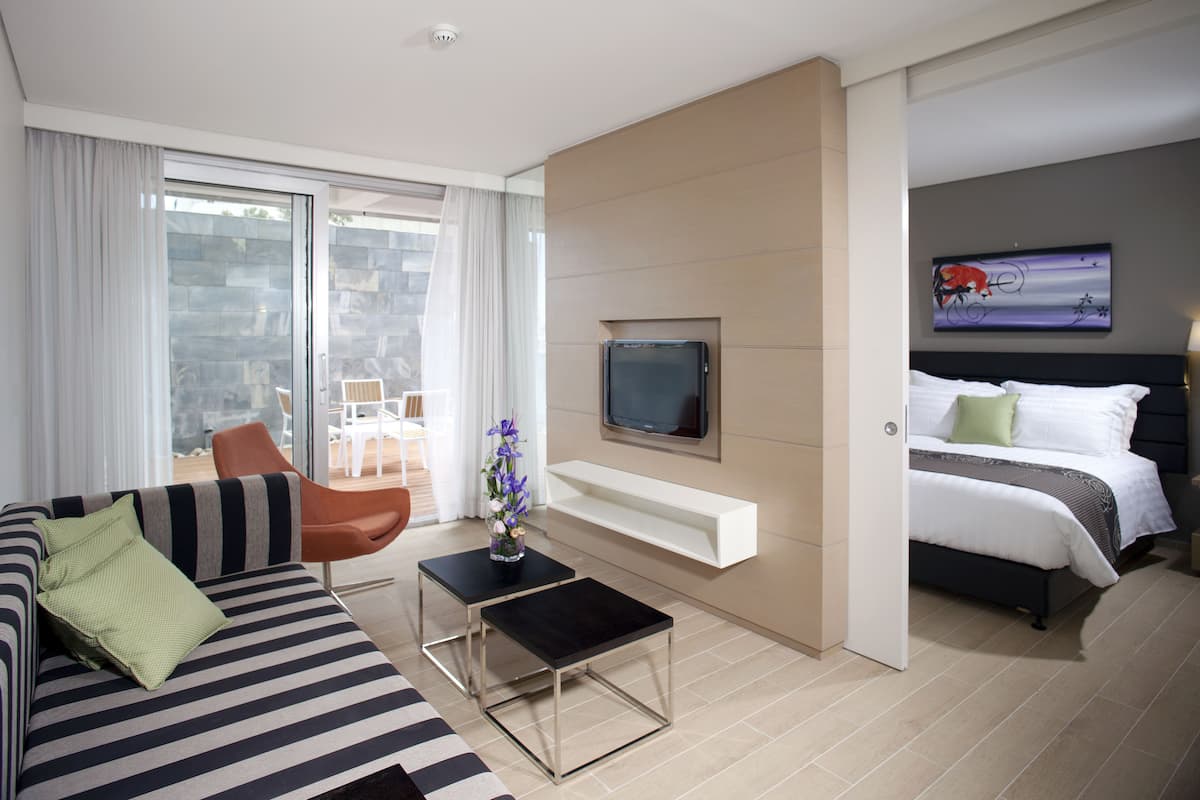 A modern hotel room with a large bed, a TV mounted on the wall, a sofa, a coffee table, and a balcony with a view of the city.