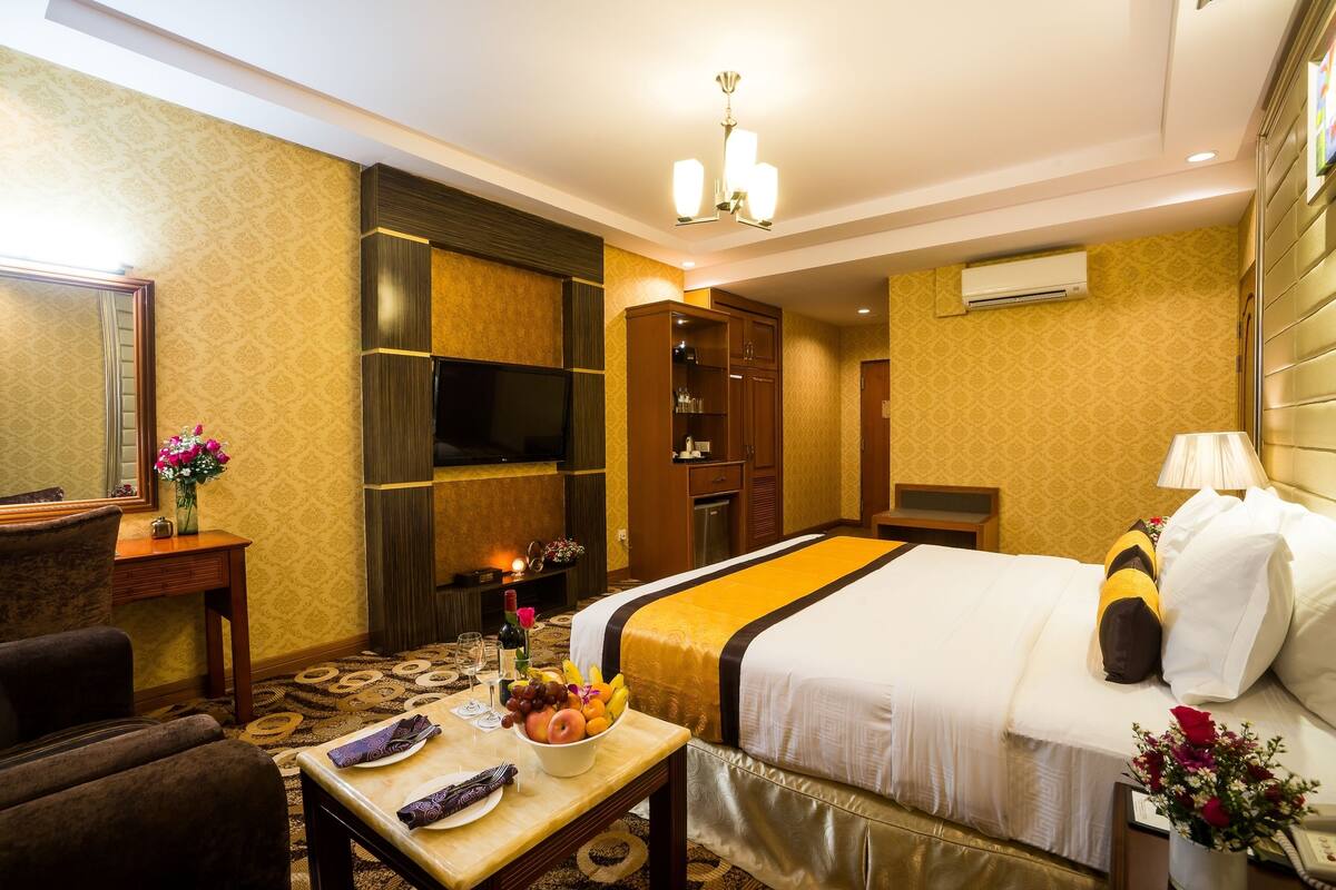 A hotel room with a large bed, a TV, a minibar, a desk with a fruit bowl, and a mirror.