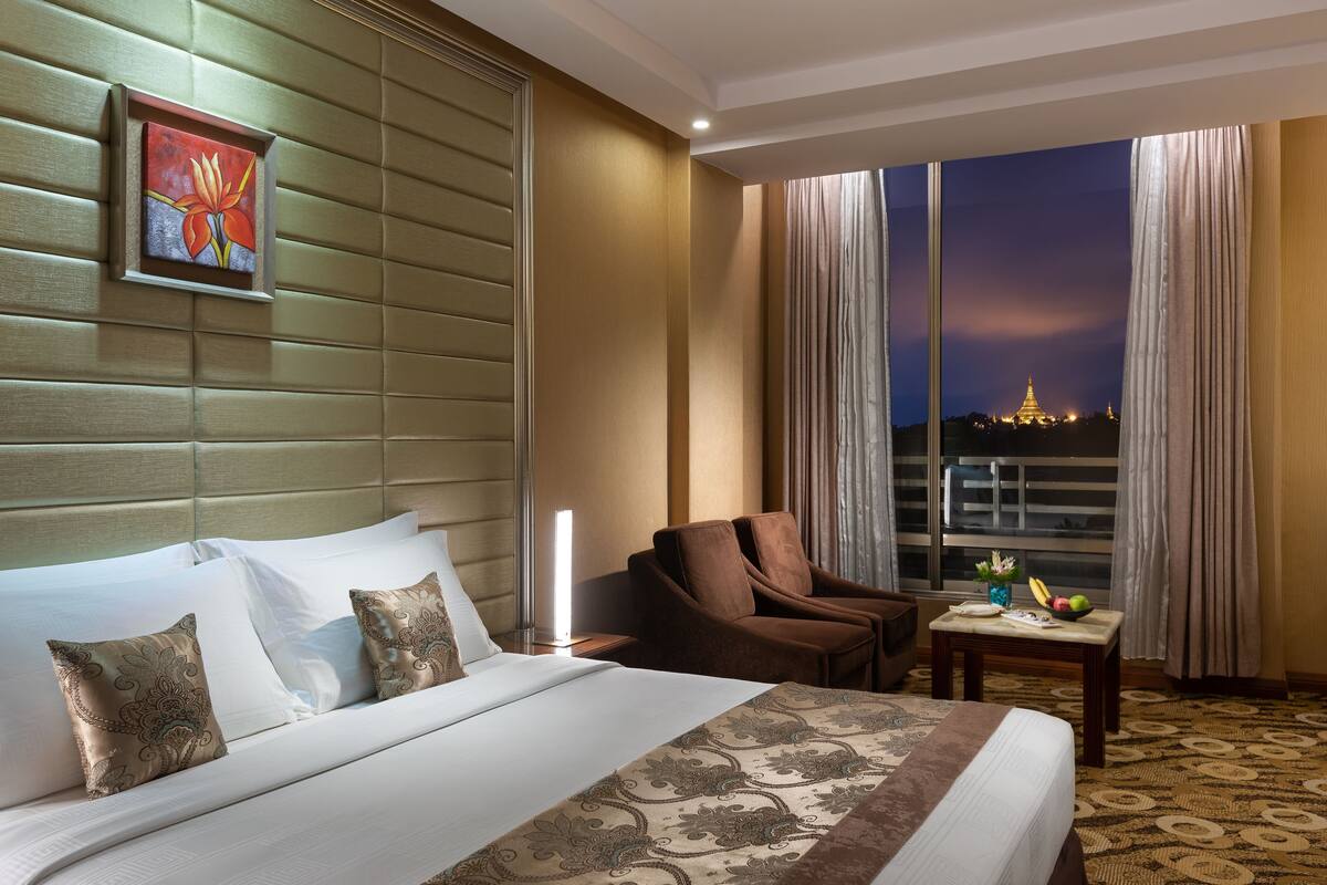 A hotel room with a bed, a chair, a table with fruits, and a view of a cityscape at night.