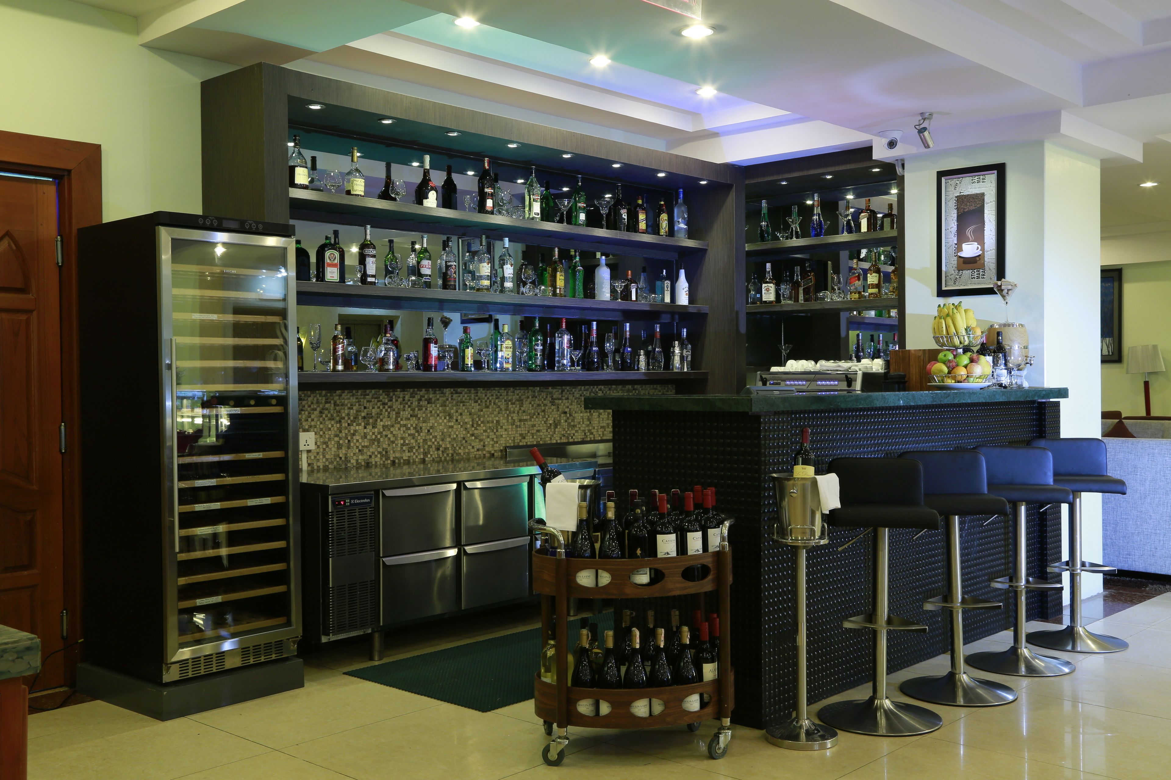 Zona de bar con estantes llenos de botellas de licor, un refrigerador y taburetes de bar.