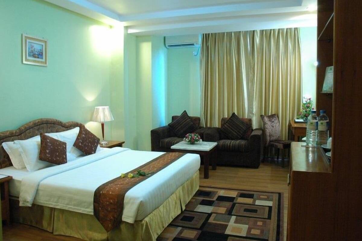 A hotel room with a bed, a sofa, a small table with a lamp, and a view of the outside through a window with curtains.