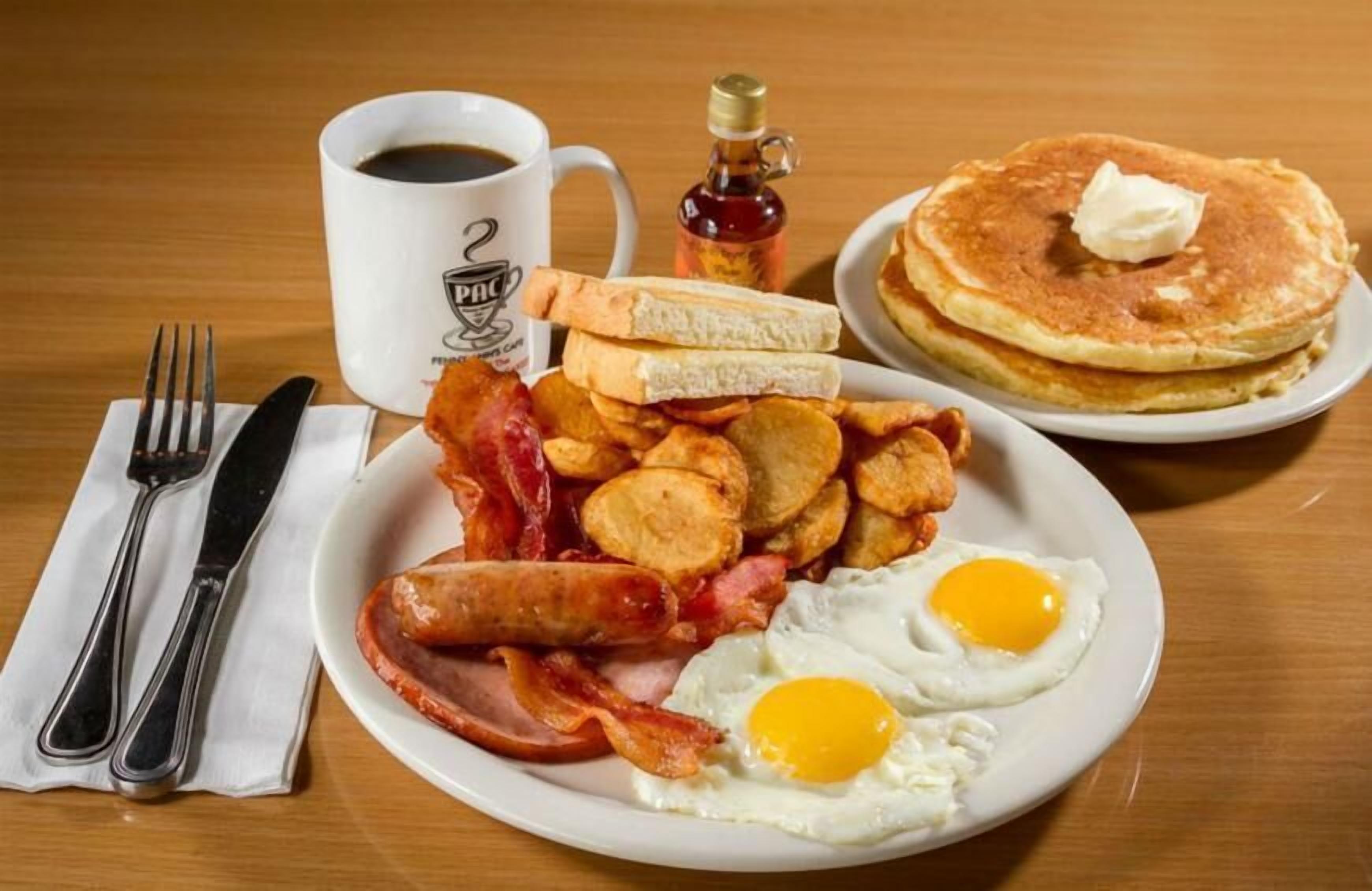 A breakfast plate with bacon, eggs, hash browns, toast, and pancakes, along with a cup of coffee and a bottle of syrup.