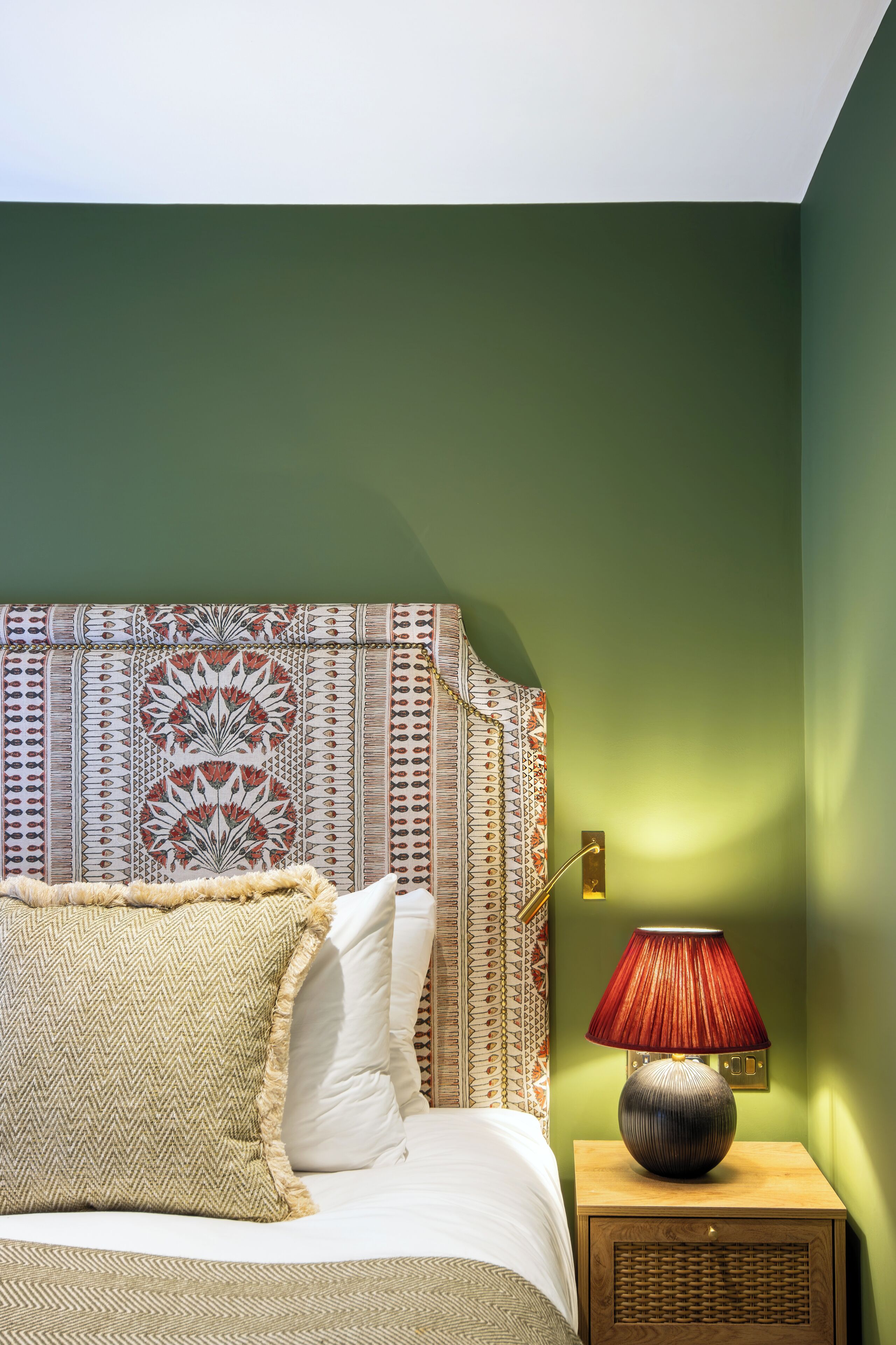 Un dormitorio con cama, almohadas, cabecera estampada, mesita de noche con lámpara y paredes verdes.