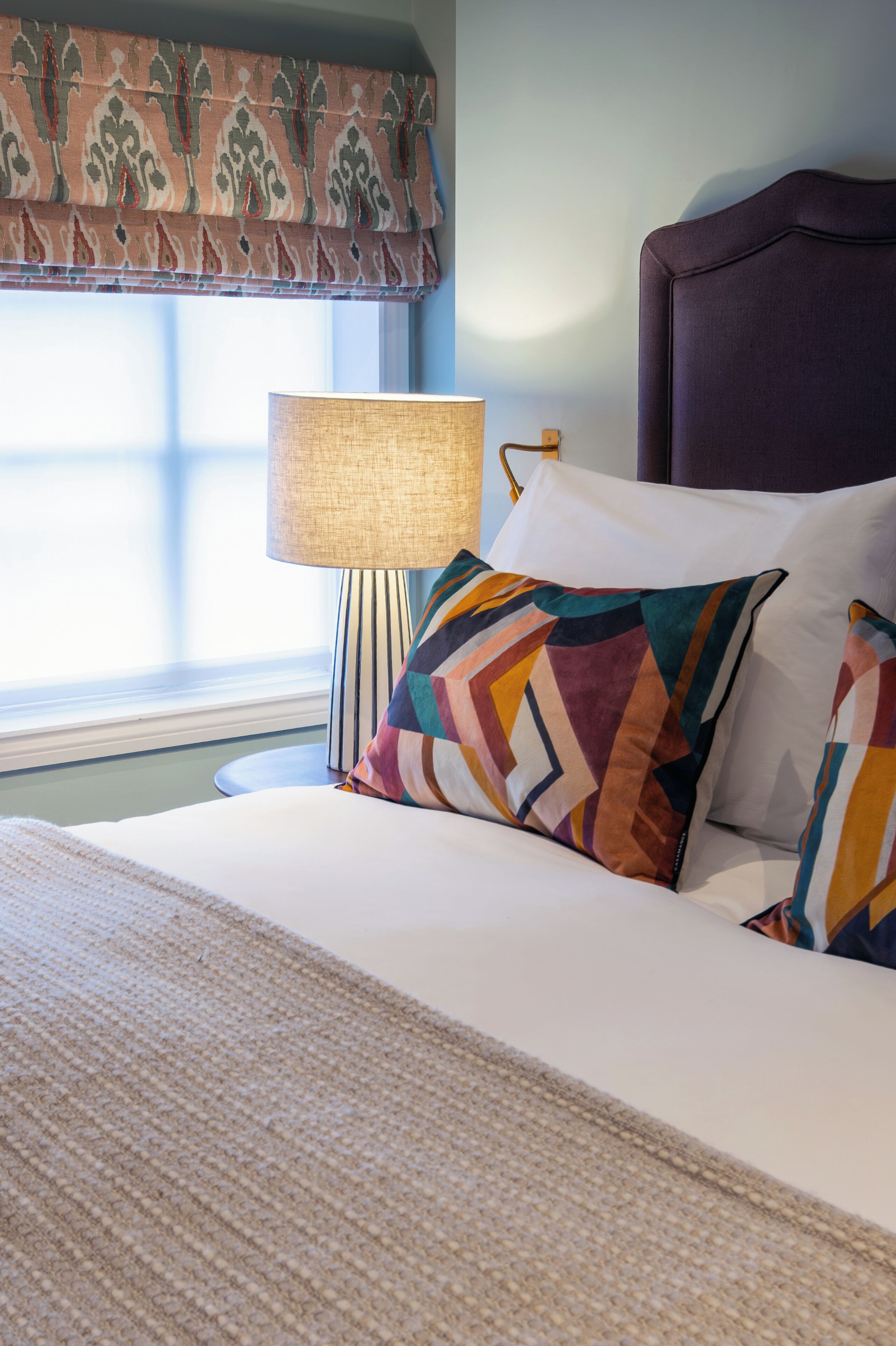 A neatly made bed with a patterned pillow, a bedside lamp, and a window with a decorative valance.