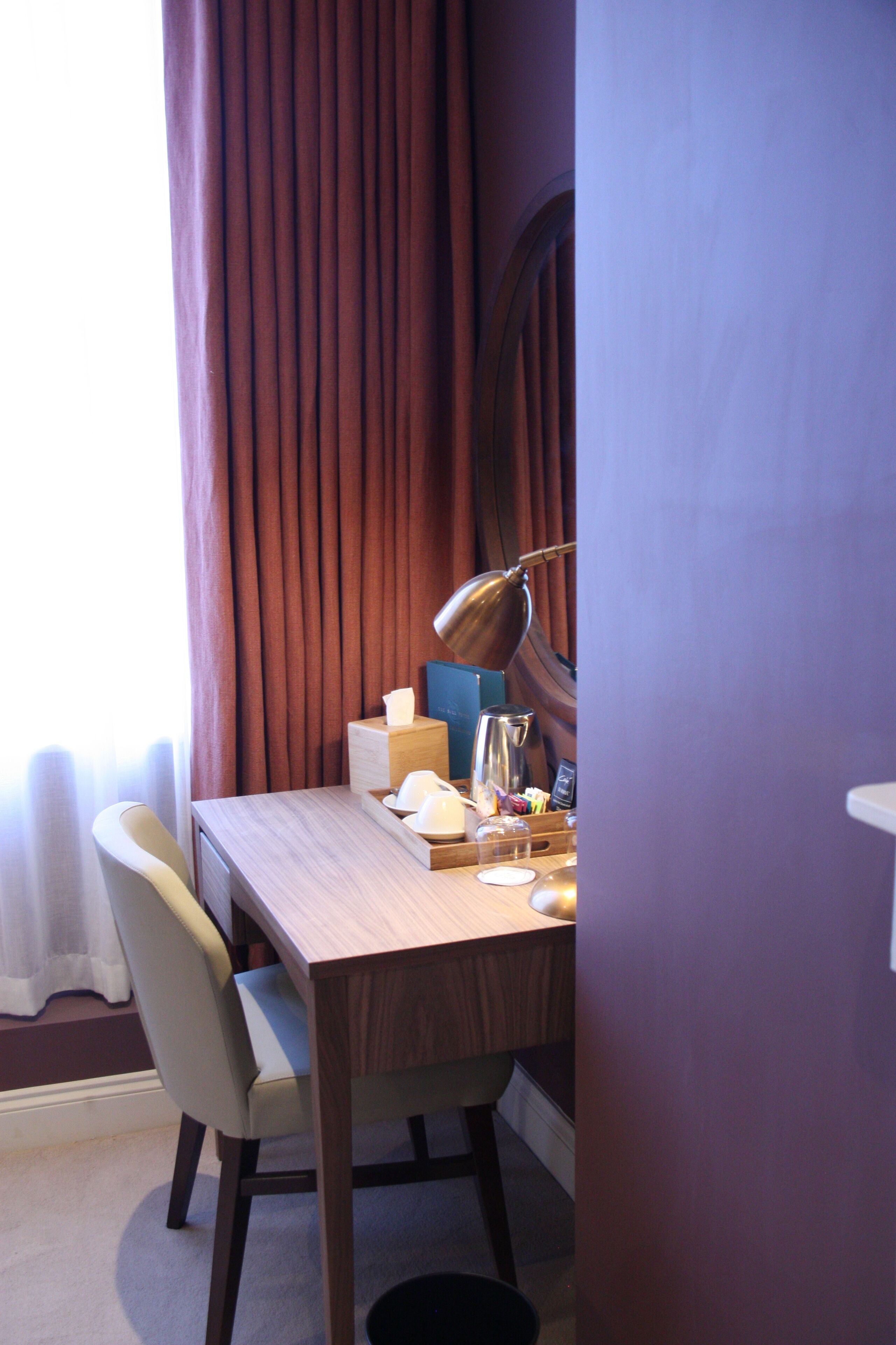 A hotel room with a wooden desk, a lamp, a chair, and a window with curtains.