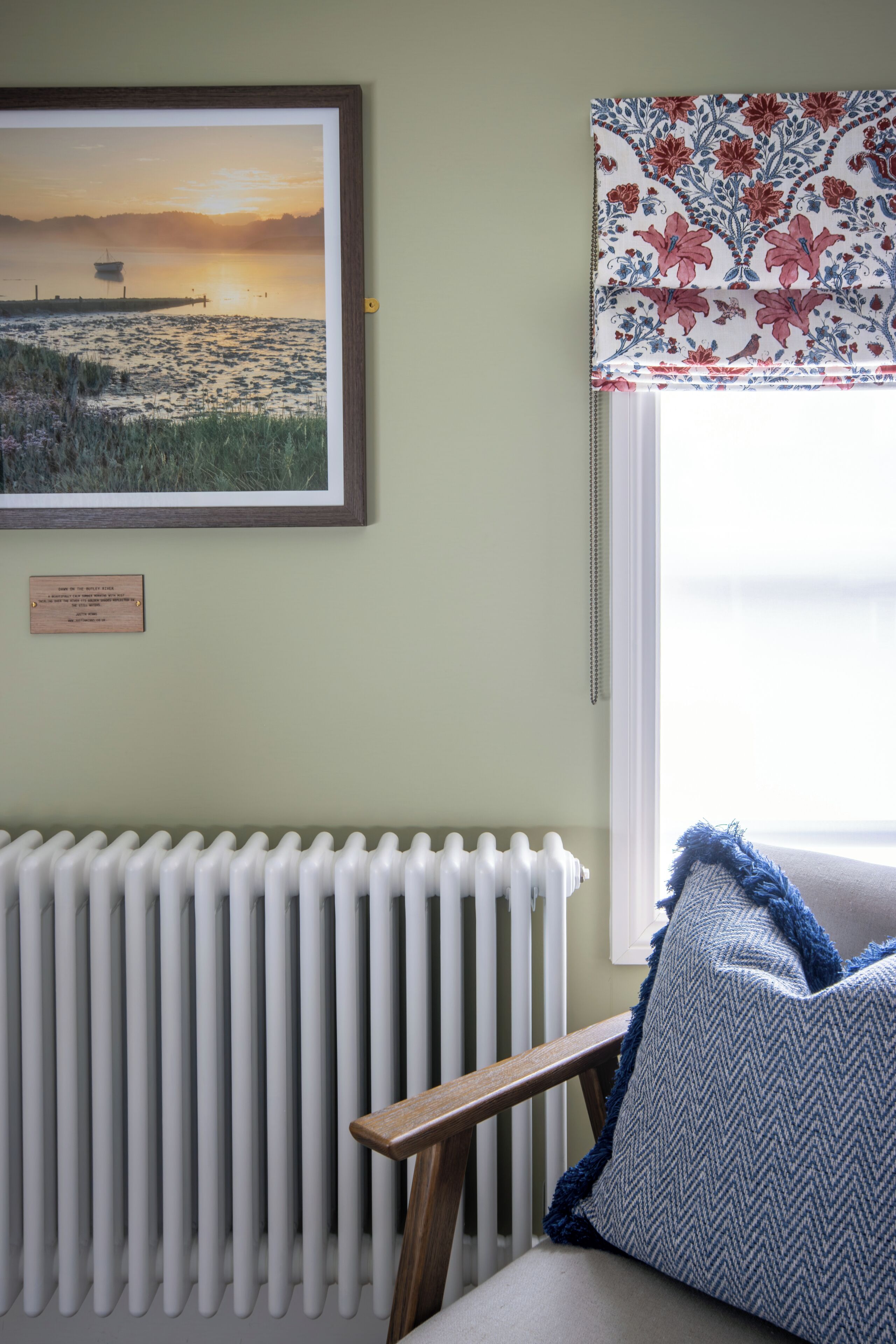 Een kamer met een radiator, een ingelijst zonsondergangschilderij en een raam met bloemenstoffering.