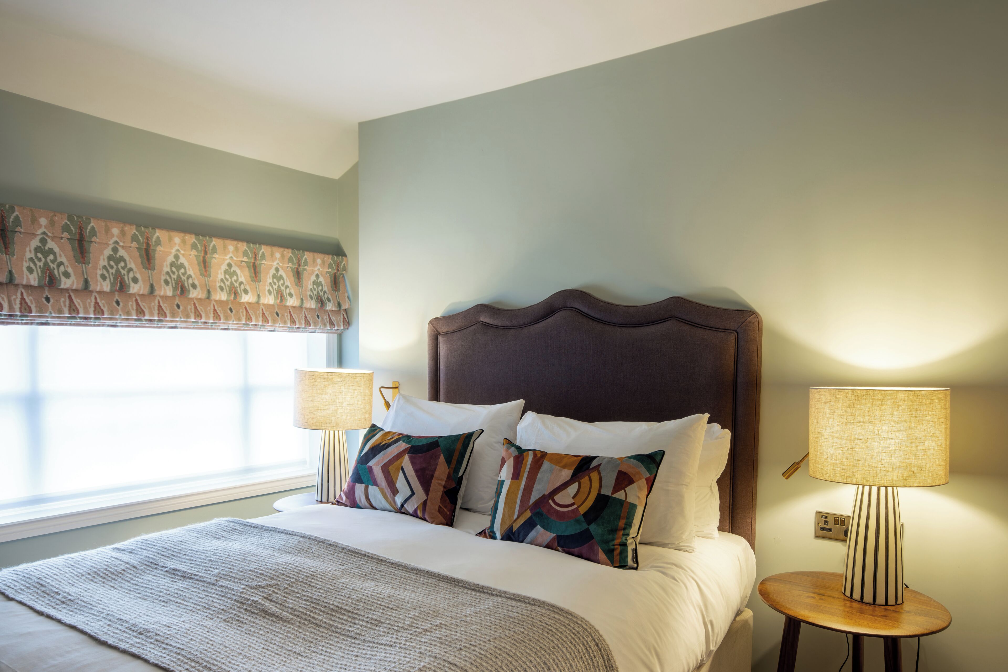 A neatly made bed with patterned pillows, a bedside lamp, and a window with a decorative valance.