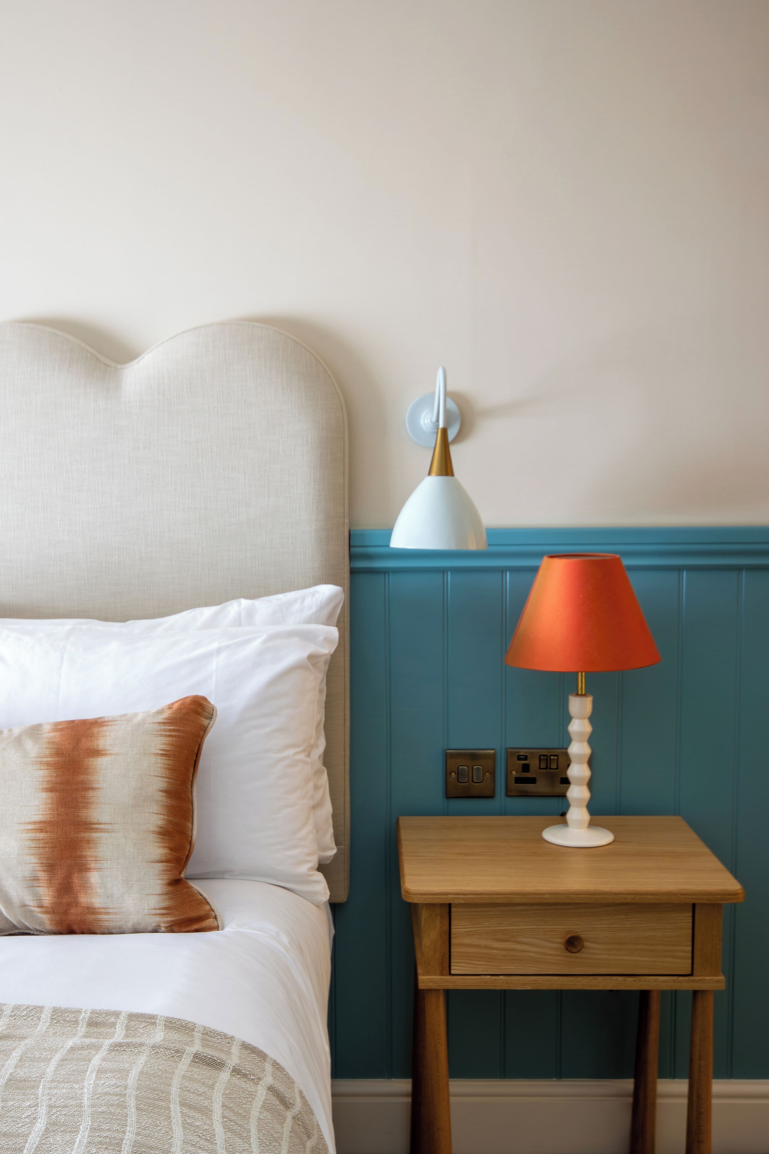 Una cama bien tendida con almohadas blancas y marrones, una mesita de noche de madera con una lámpara y un interruptor de pared.
