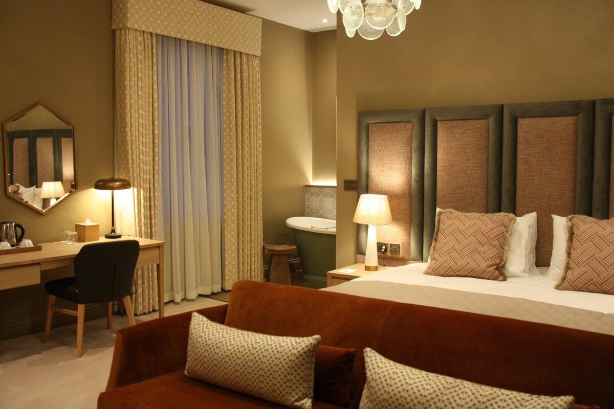 A hotel room with a bed, a desk, a chair, a couch, a mirror, a lamp, and a chandelier.