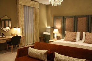 Hotellihuone, jossa on sänky, työpöytä, tuoli, sohva, peili, lamppu ja kattokruunu.