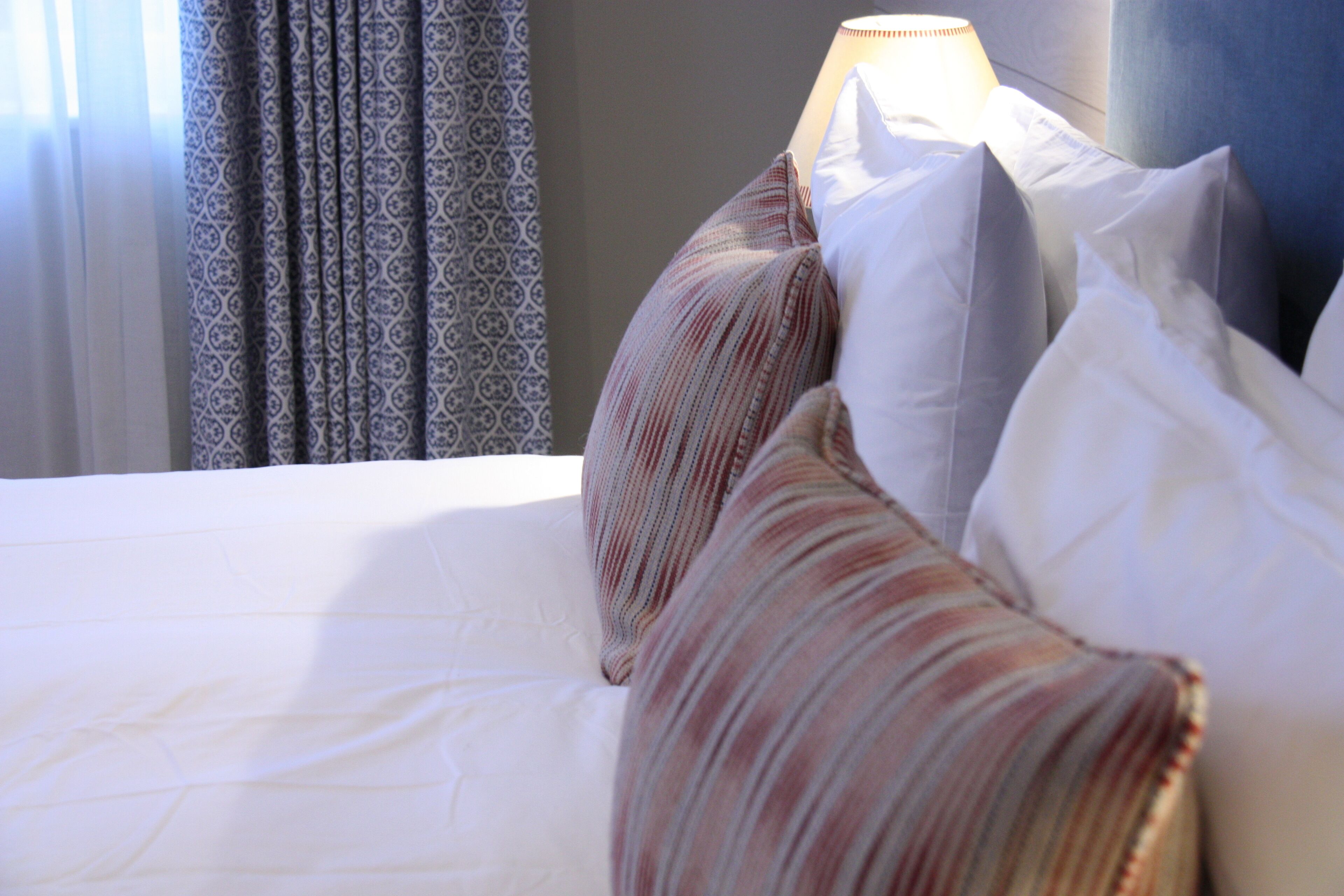 A neatly made bed with white bedding and striped pillows, a lamp on the side, and patterned curtains in the background.