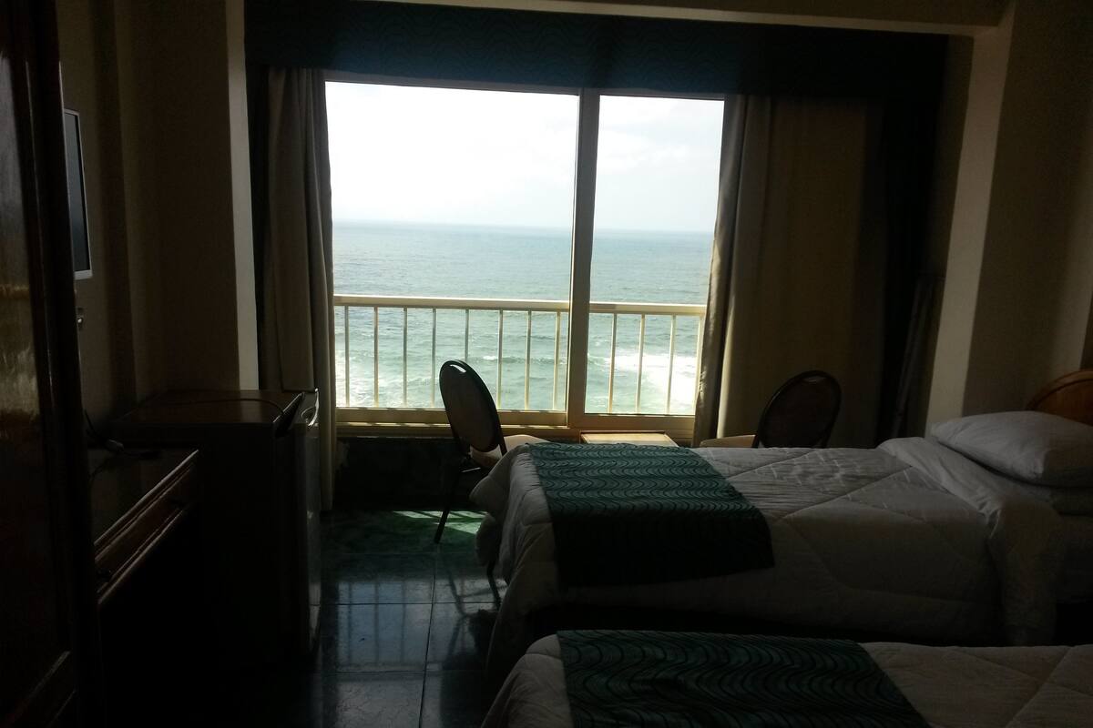 A hotel room with two beds, a chair, a desk, and a view of the ocean through large windows.