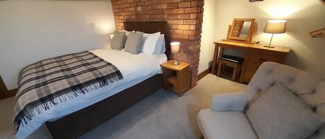 A bedroom with a brick fireplace, a bed with a plaid blanket, a bedside table with a lamp, and a chair.