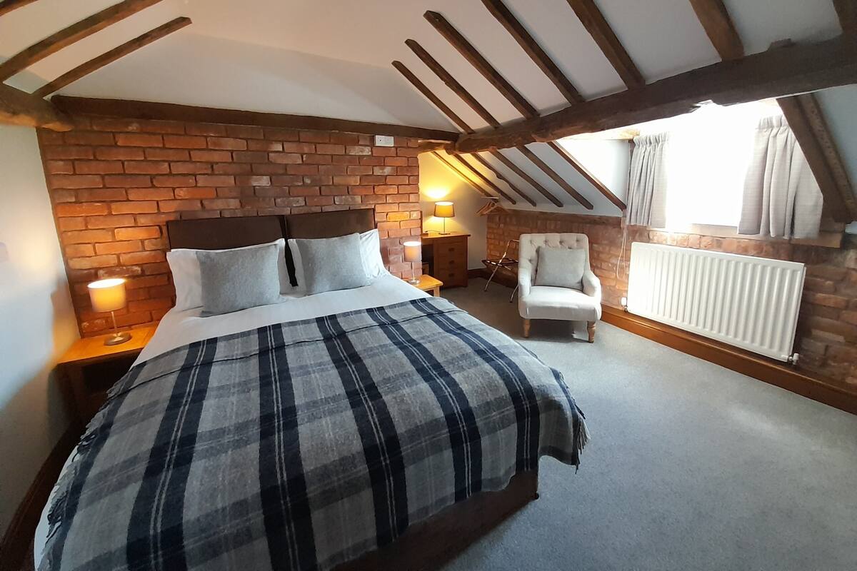 Ein Schlafzimmer mit Backsteinwand, einem Bett mit karierter Decke, einem weißen Sessel und einem Fenster mit Vorhängen.