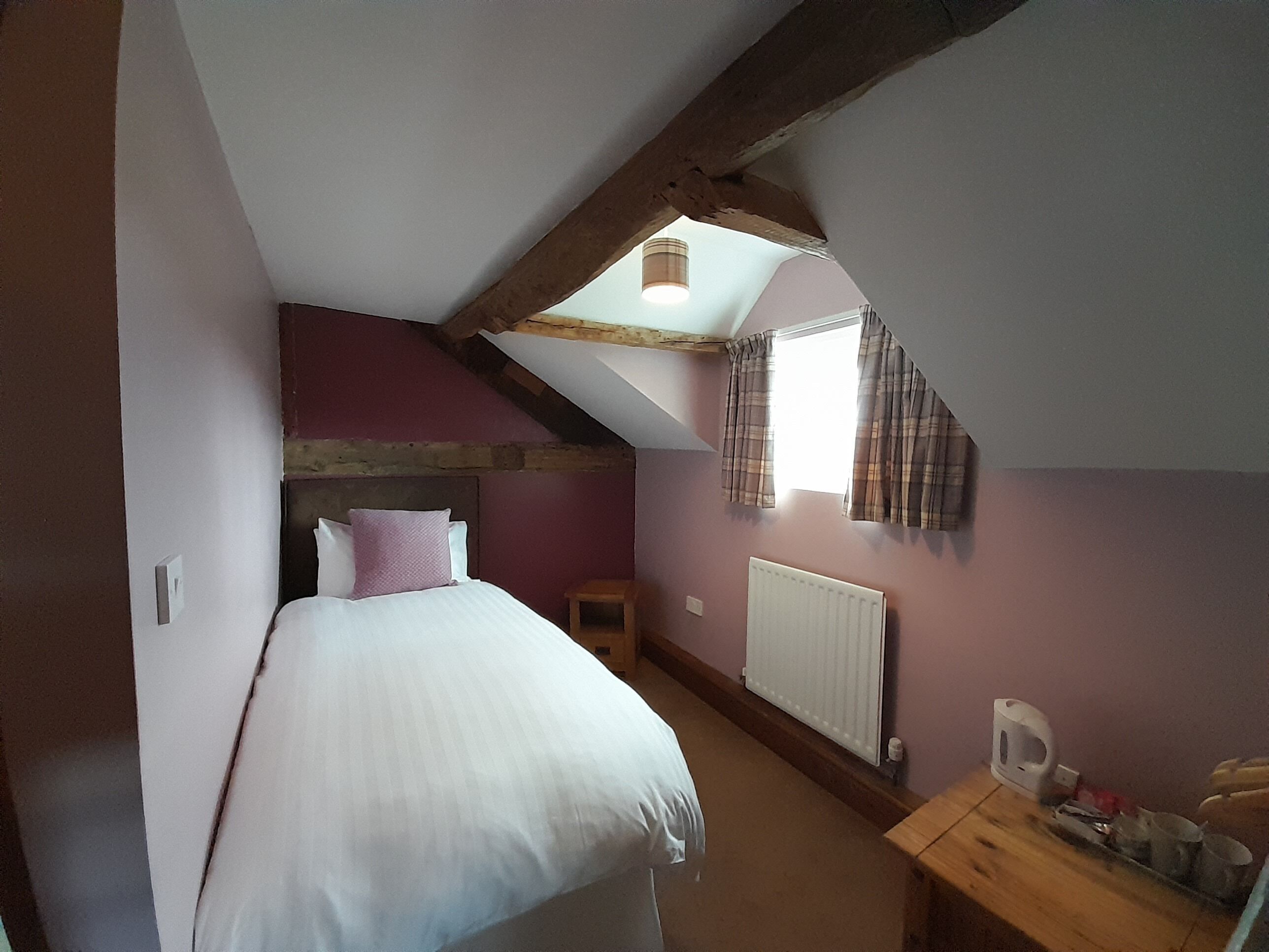 A bedroom with a sloped ceiling, a bed with white bedding, a small wooden nightstand, a radiator, and a window with curtains.