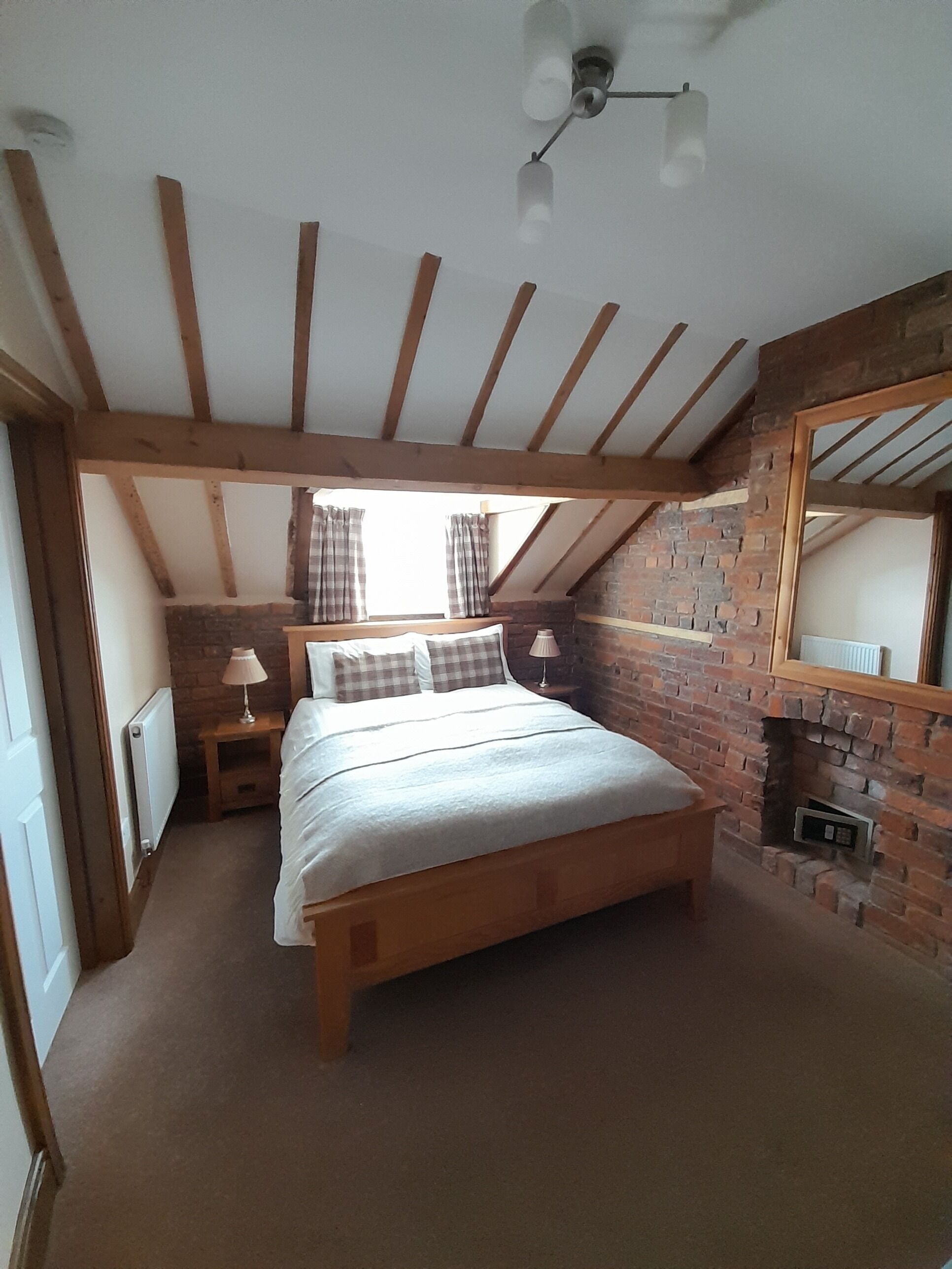 A bedroom with a wooden ceiling, a brick wall, a bed with pillows, two bedside tables with lamps, and a window with curtains.