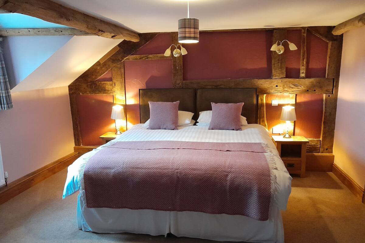 Ein Schlafzimmer mit einem großen Bett, Holzbalken, zwei Nachttischlampen und einer geneigten Decke.