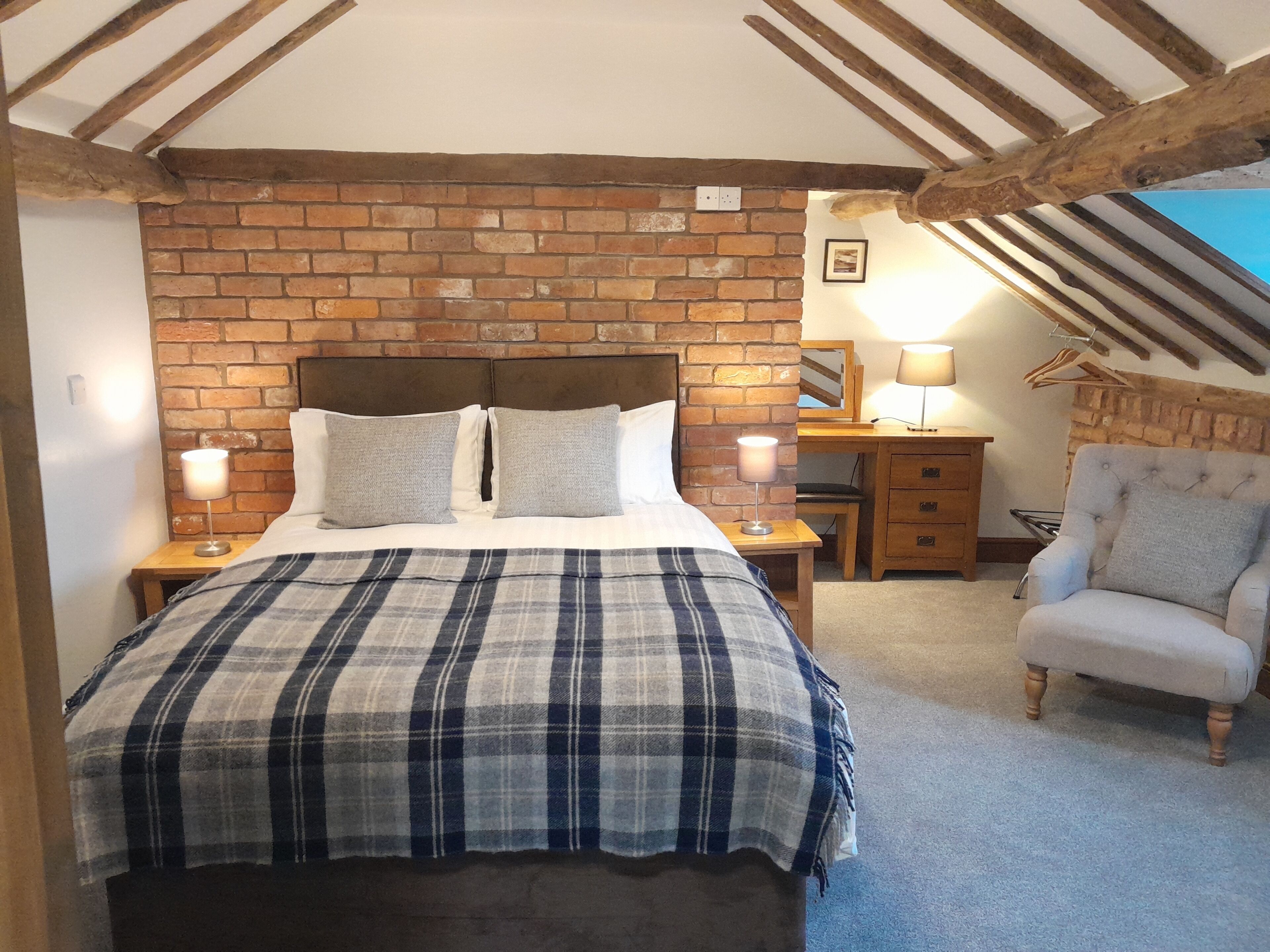 Een slaapkamer met een bakstenen muur, houten balken, een bed met een geruite deken, bedlampen, een houten nachtkastje, een stoel en een bureau met een lamp.