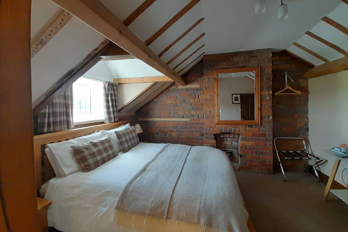 Ein Schlafzimmer mit geneigter Decke, einem Kamin aus Ziegelsteinen, einem Bett mit Karo-Bettwäsche, einem Holzstuhl und einem Spiegel an der Wand.