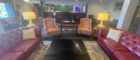 A living room with two red leather sofas, a black coffee table, and patterned armchairs. There are lamps on side tables, a large rug, and a television in the background.