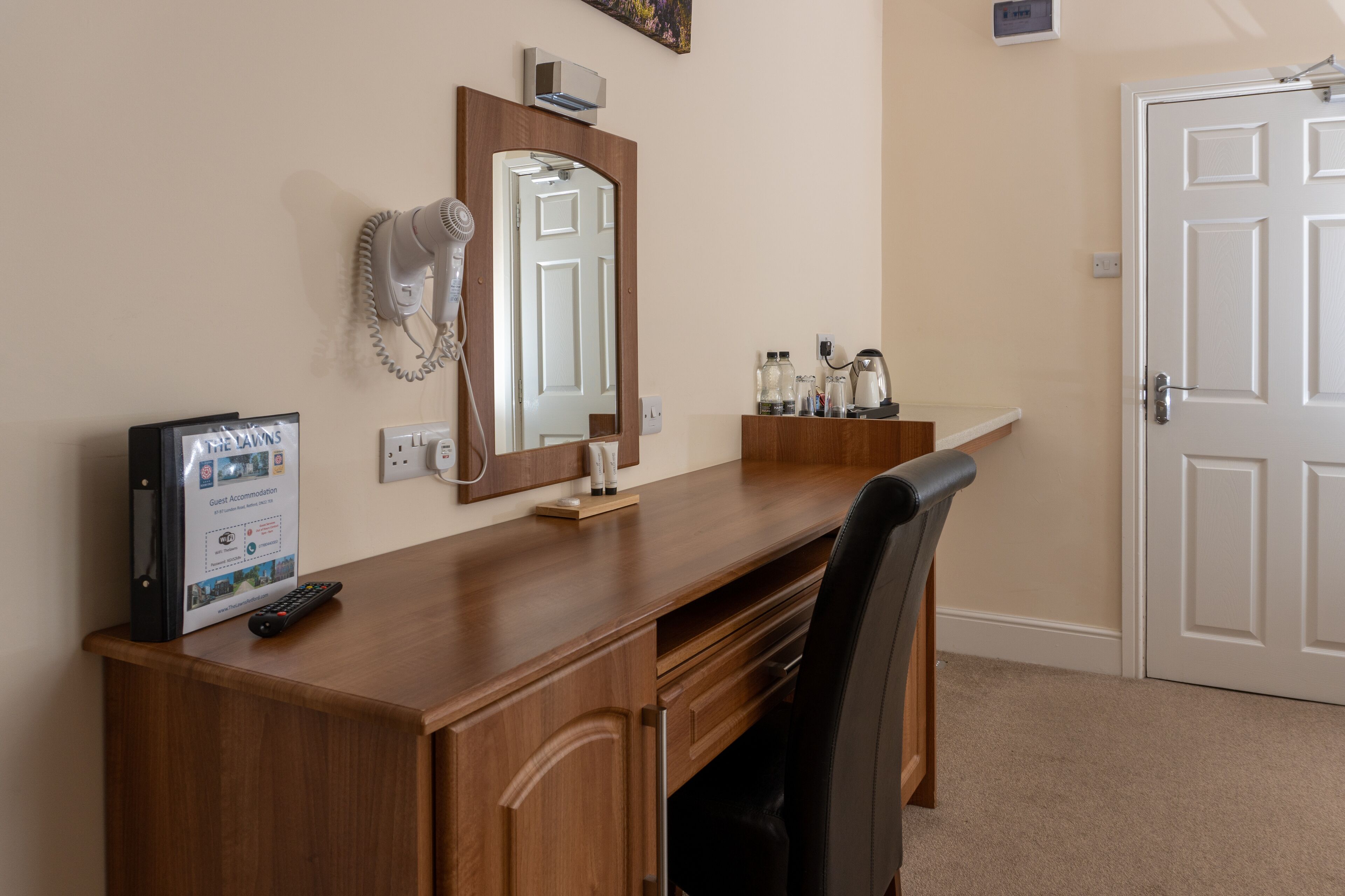 A hotel room with a wooden desk, a hairdryer, a mirror, a framed sign, and a door.