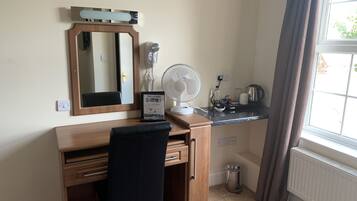 Habitación de hotel con escritorio de madera, una silla negra, un espejo, un ventilador, un hervidor y un radiador.