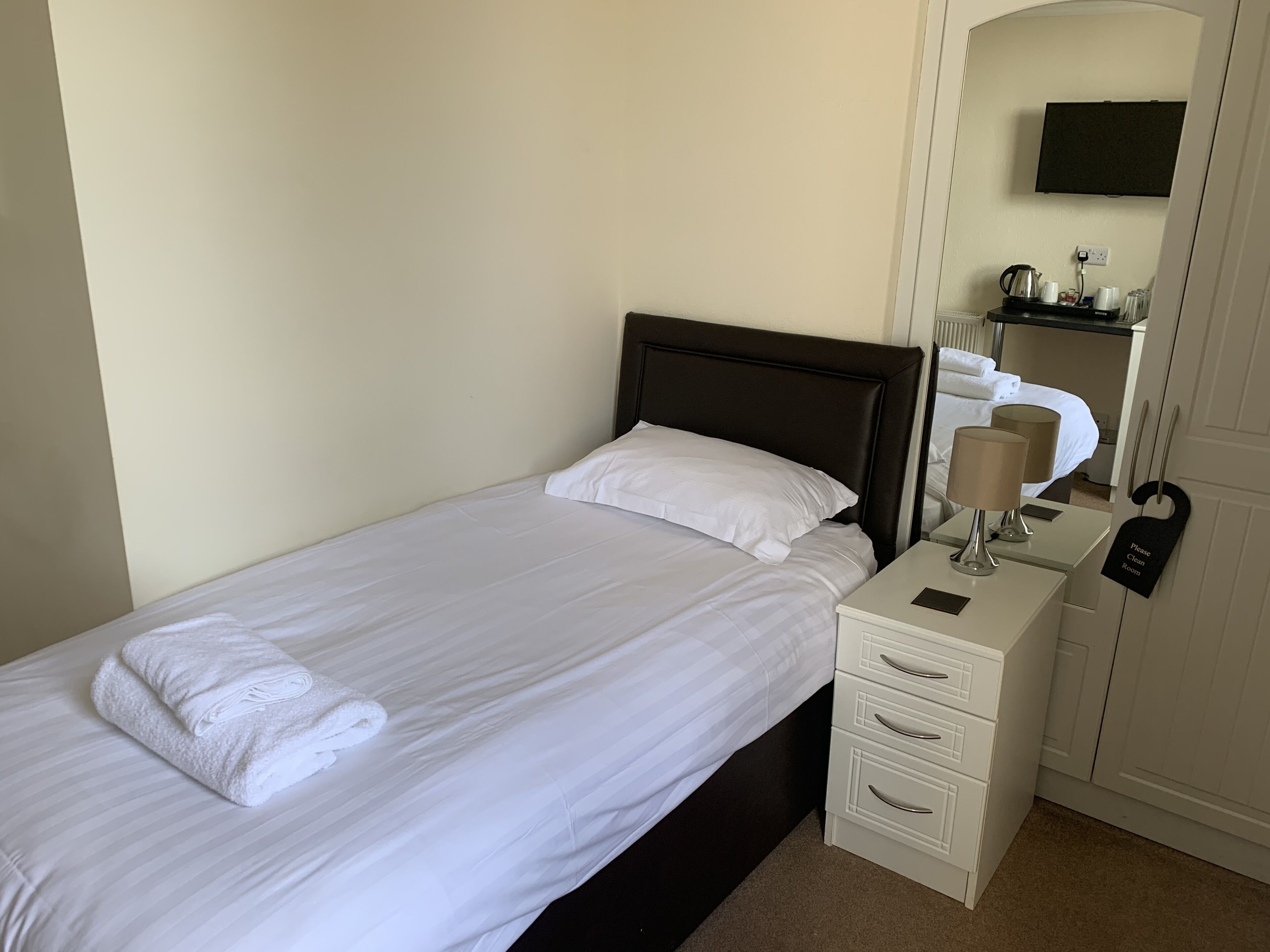 A neatly made bed with white linens and a folded towel, a bedside table with a lamp, a small shelf with toiletries, and a "Please Clean Room" sign.
