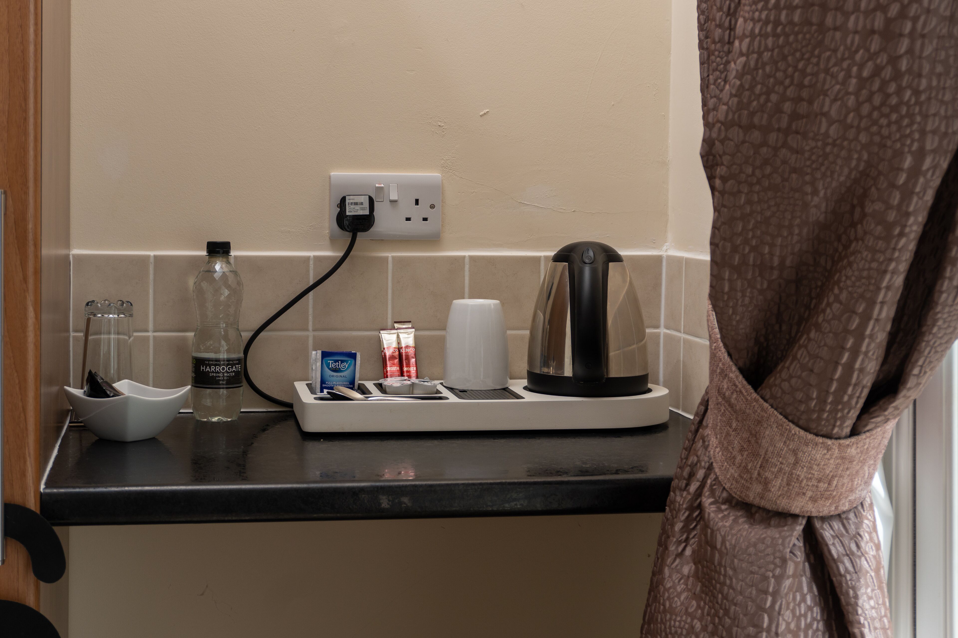 A kitchen counter with a coffee maker, a bottle of Harrogate water, a bottle of Tetley tea, a white cup, and a small bowl.