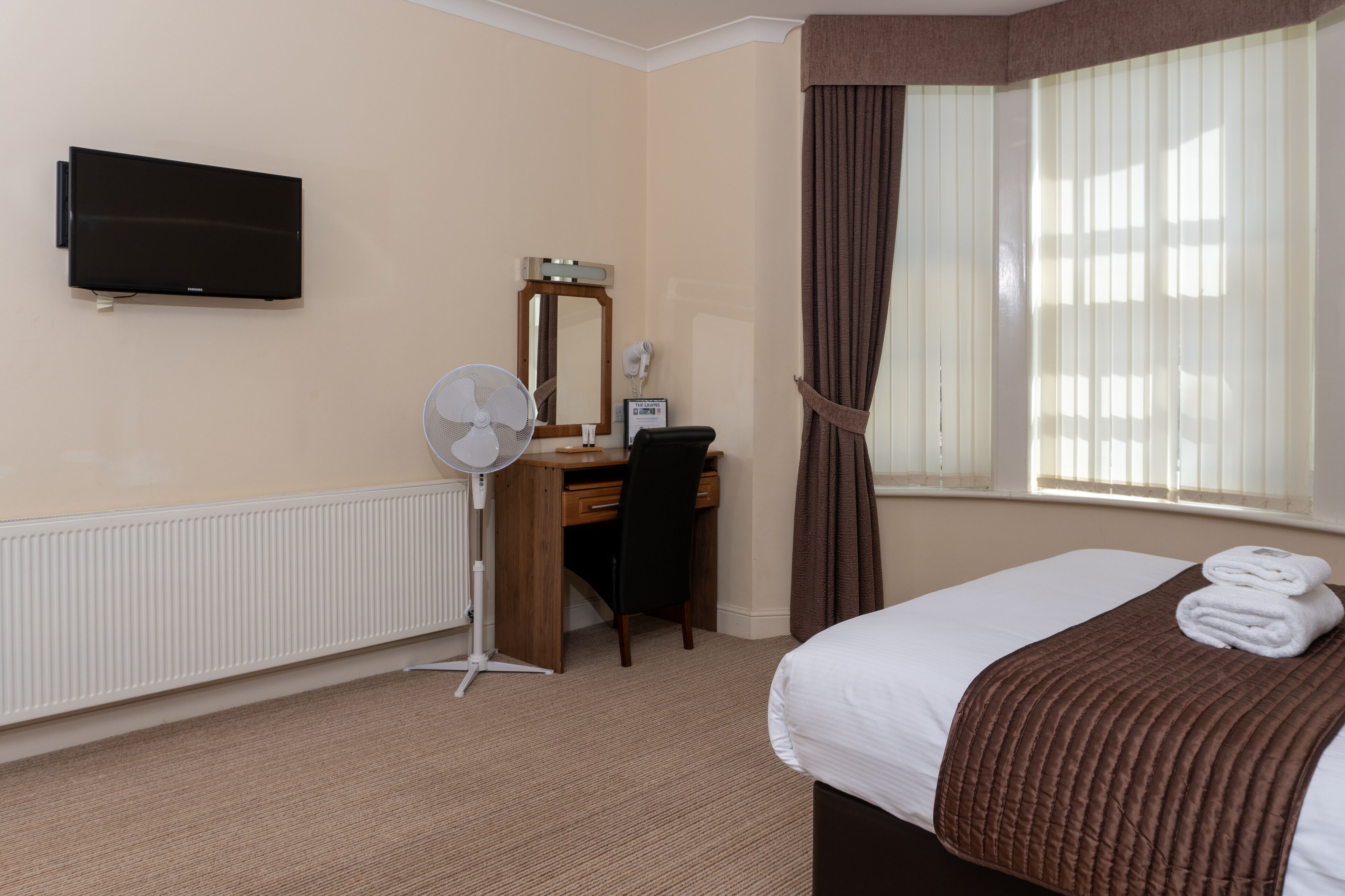A hotel room with a bed, a desk with a chair, a flat-screen TV, a standing fan, a mirror, and a window with curtains.