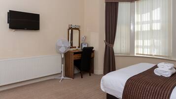 A hotel room with a bed, a desk with a chair, a flat-screen TV, a standing fan, a mirror, and a window with curtains.