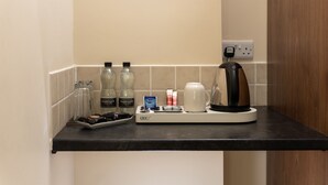 Una kitchenette con cafetera, dos botellas de Harrogate, una copa, un plato con aperitivos y una cápsula de café.