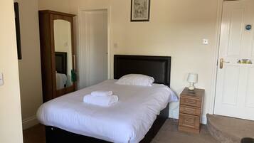 A neatly made bed with white linens and a folded towel, a wooden nightstand with a lamp, a door with a caution sign, and a framed picture on the wall.