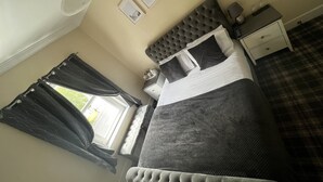 Une chambre à coucher avec une tête de lit capitonnée, un lit avec une literie aux couleurs sombres, une table de chevet, une lampe et une fenêtre avec des rideaux.